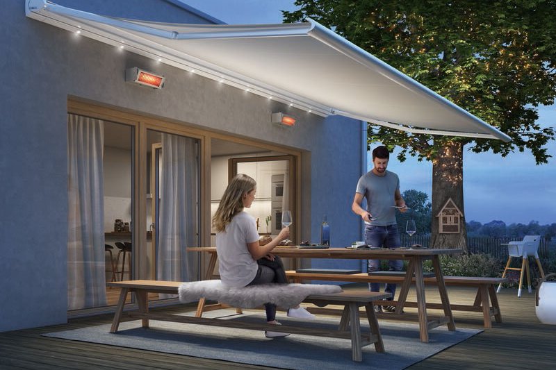 A couple enjoying an evening on a terrace under a retractable awning, with a tree illuminated in the background. A couple enjoying an evening on a terrace under a retractable awning, with a tree illuminated in the background.