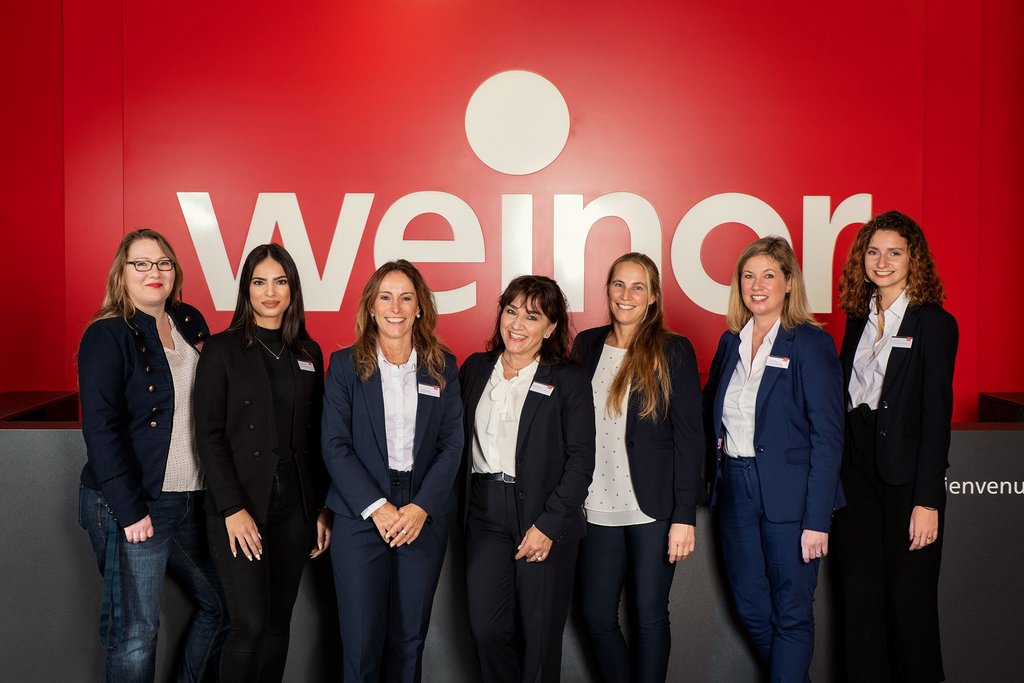 A group of seven women in professional attire stands in front of a red wall with the word "weinor" displayed. A group of seven women in professional attire stands in front of a red wall with the word "weinor" displayed.